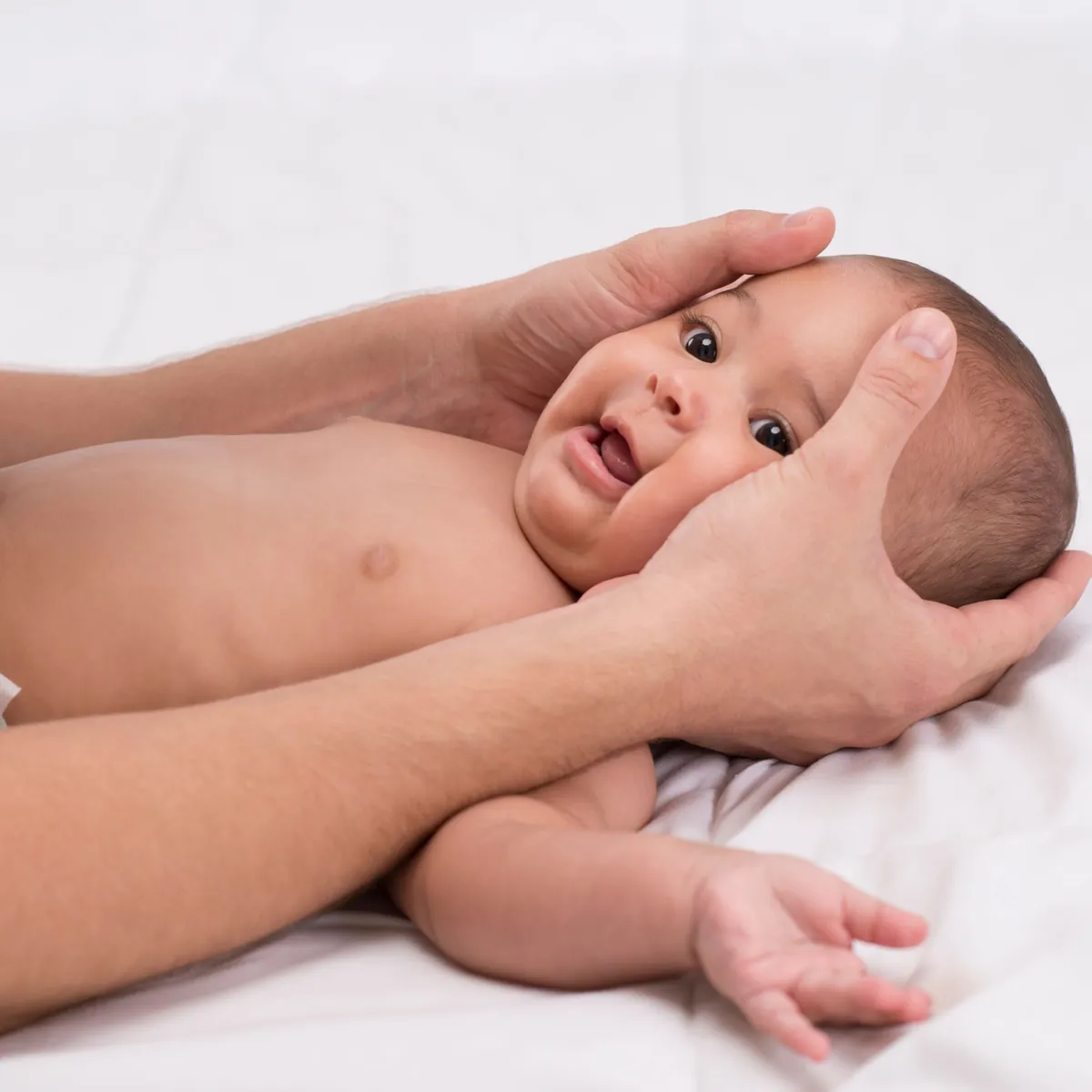 Ein Baby liegt auf dem Rücken und schaut lächelnd zur Seite, zwei Hände halten behutsam seinen Kopf