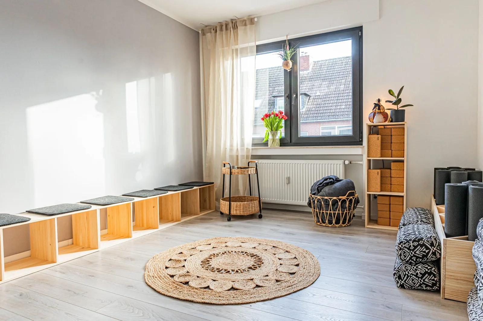 Sonniger Yogaraum mit runder Matte, Sitzbank aus Holz, Korkblöcken und Decken im Korb