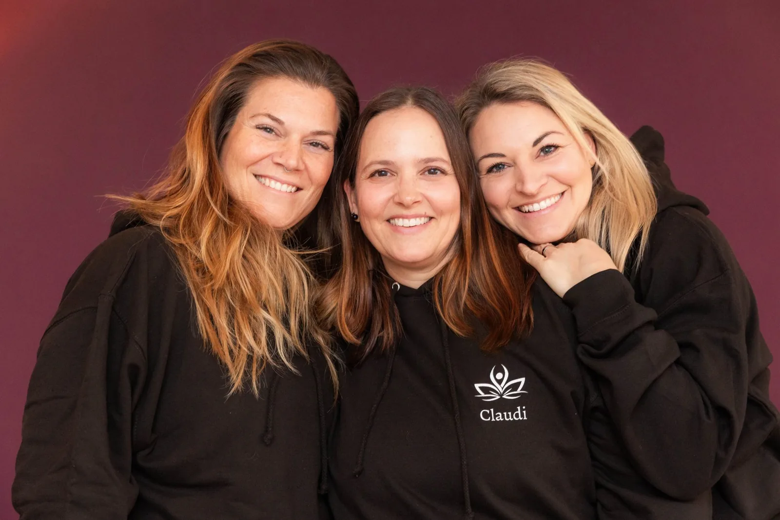 Drei Yogalehrerinnen in dunklen Hoodies lachen dicht nebeneinander vor beerenrotem Hintergrund