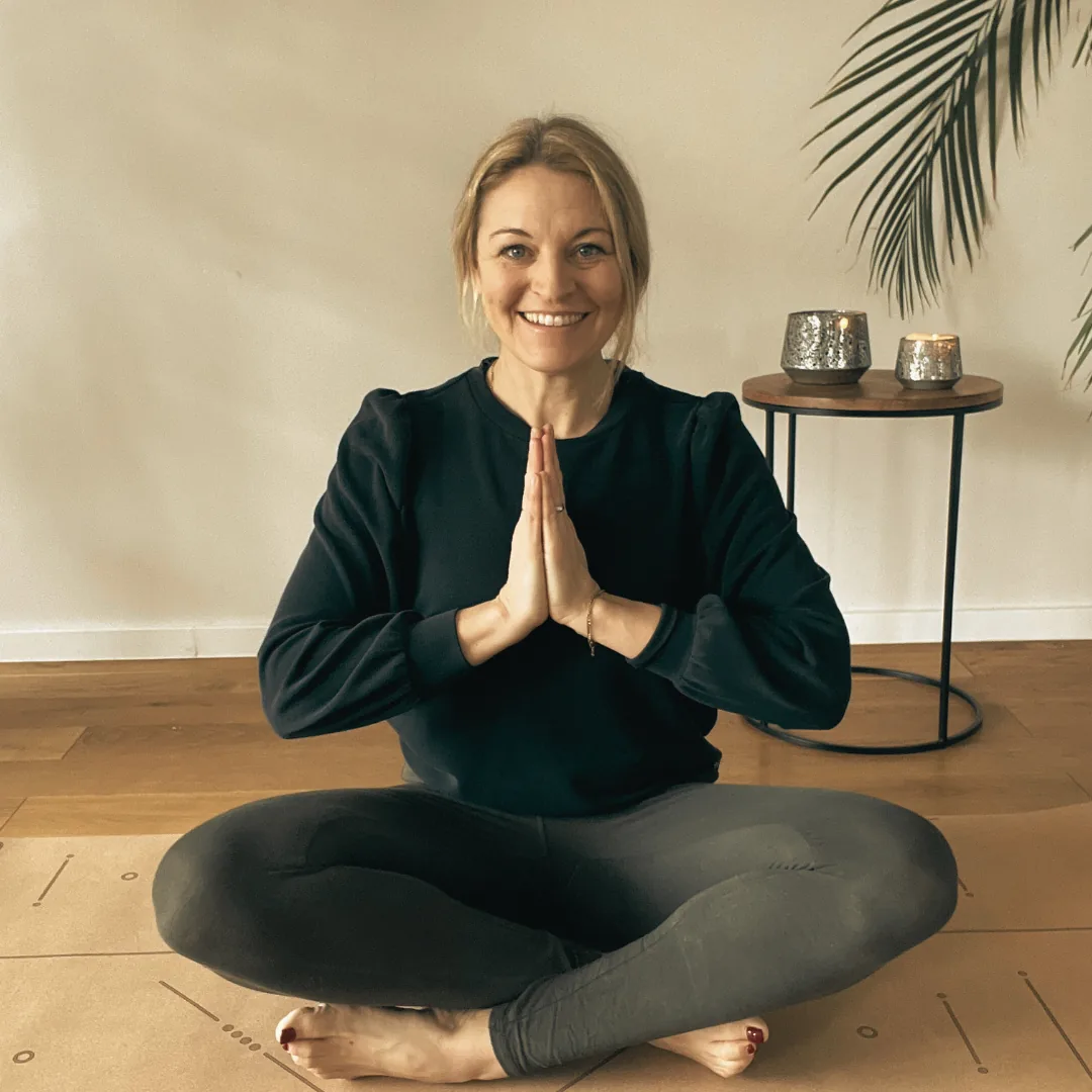 Yogalehrerin sitzt lächelnd im Schneidersitz auf einer Matte, die Hände vor der Brust