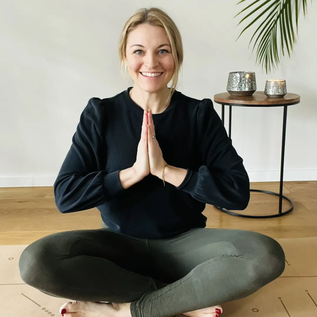 Chrissi, Gründerin von YogaPänz, sitzt lächelnd im Schneidersitz auf einer Yogamatte im Yogastudio Pulheim, die Hände vor der Brust zusammengelegt